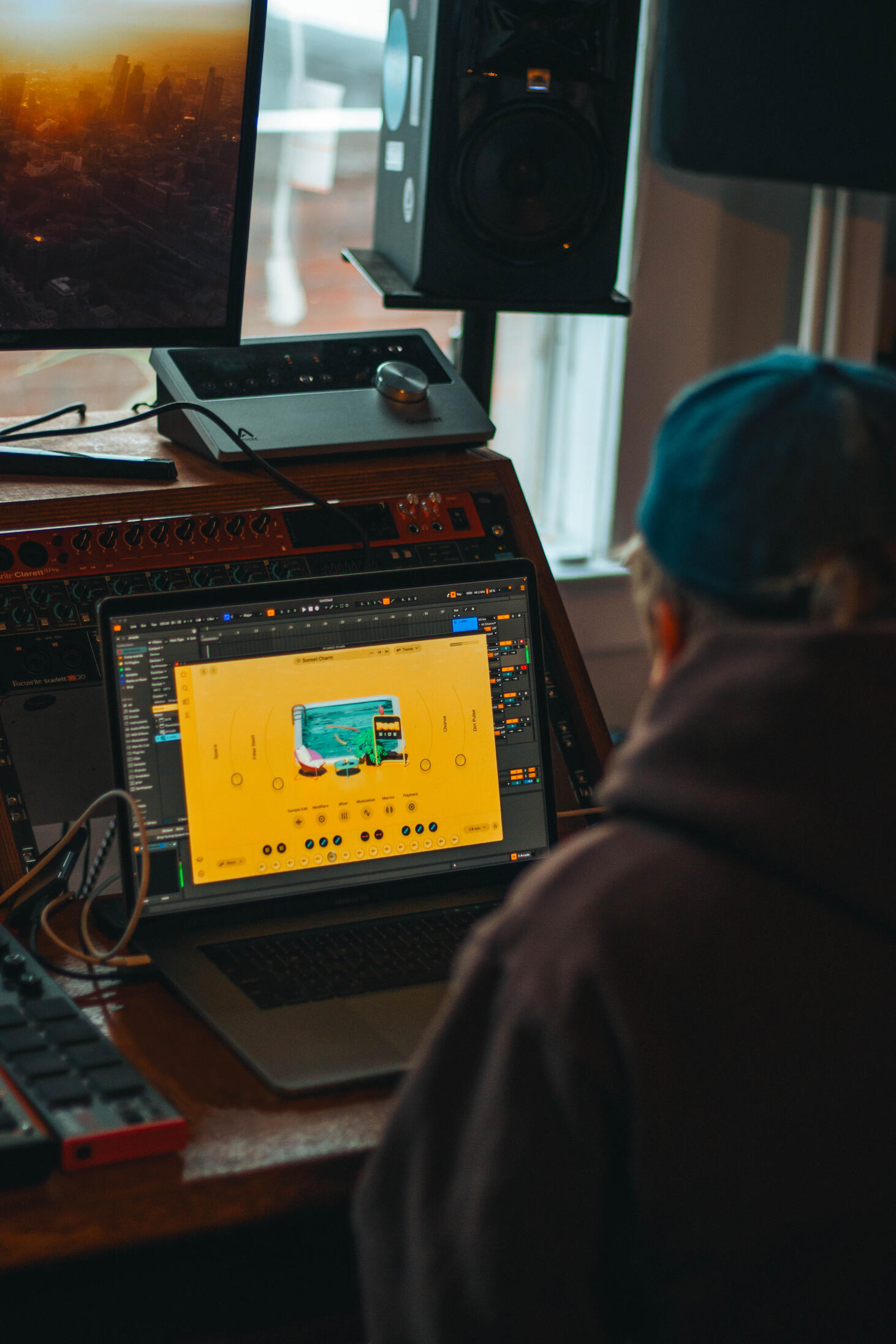 A man in a denim blue baseball cap sitting in front of a laptop and mixing desk with an Ableton plugin designed for making new sounds for music on the screen.