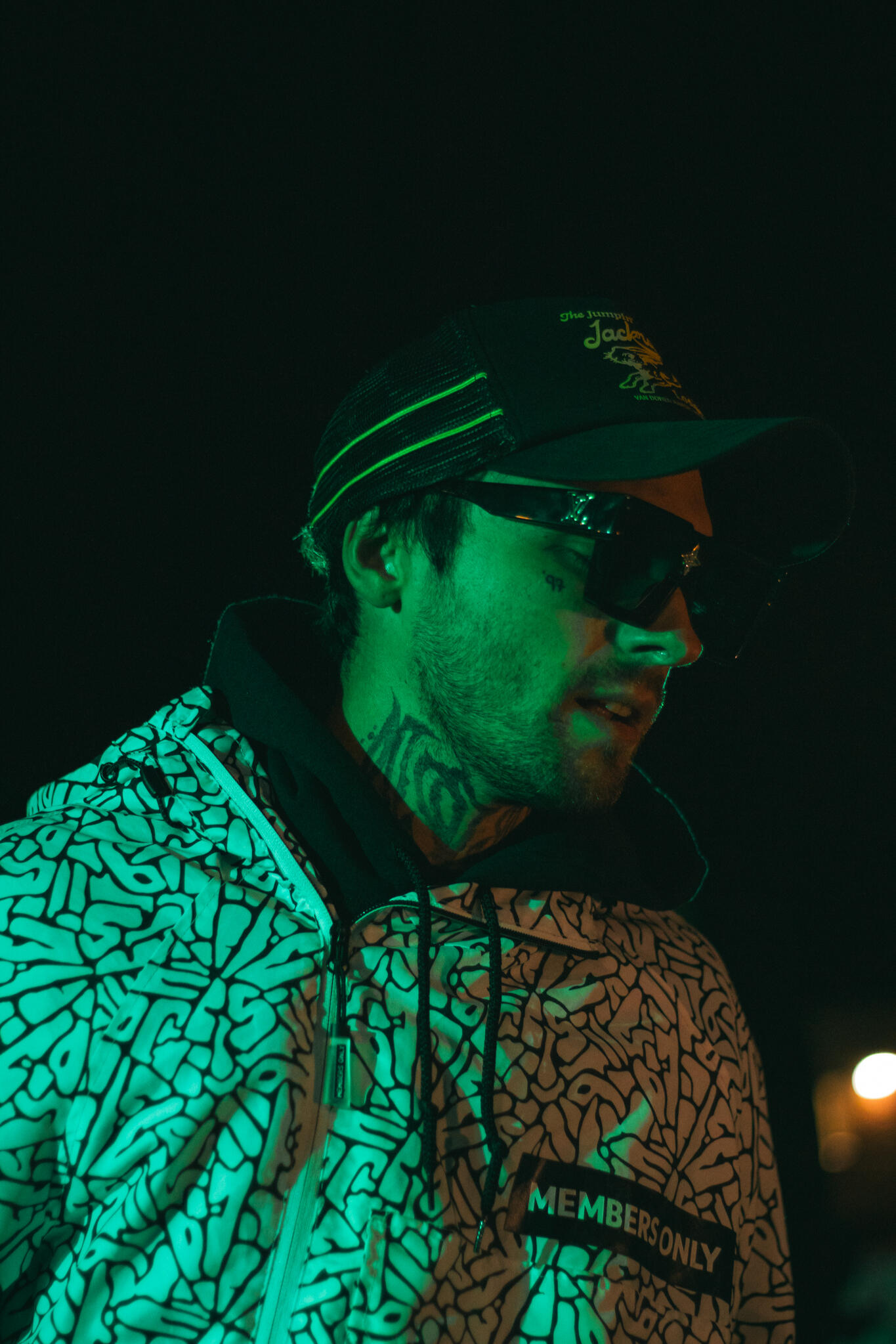 A man wearing a hat and Members Only jacket stands on stage in a dimly lit environment, creating a mysterious atmosphere while he performs rap music.