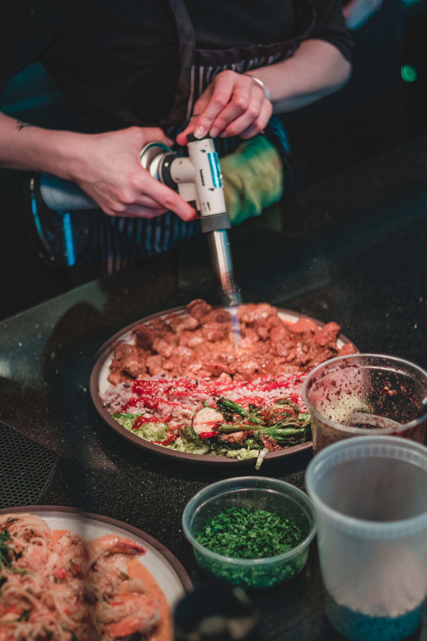 A chef using a food torch to finish of his Korean Italian infusion dish on a nice countertop with other ingredients surrounding it.