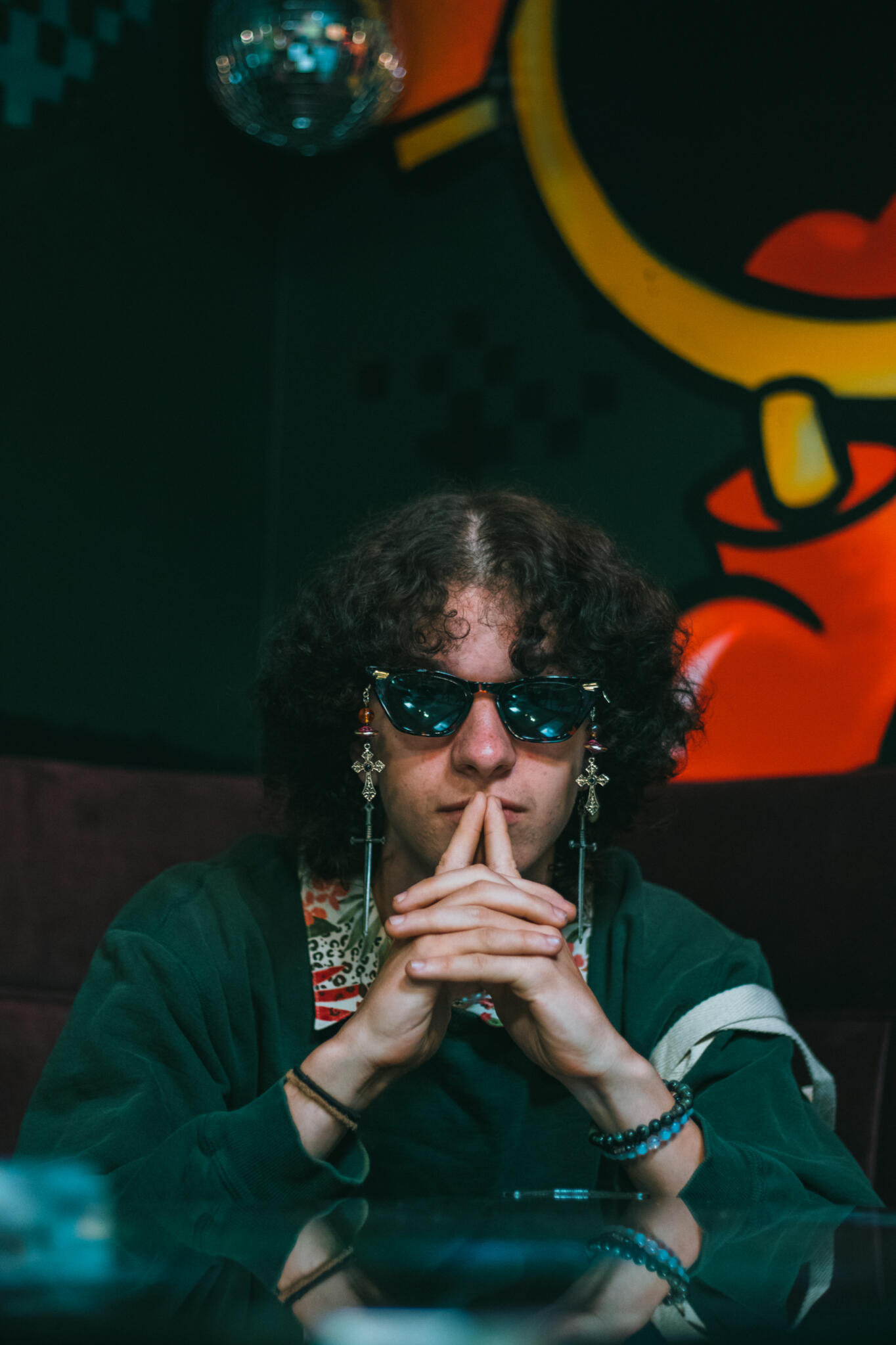 A man with curly hair and sunglasses is sitting at a table, exuding a laid-back vibe in a social environment.