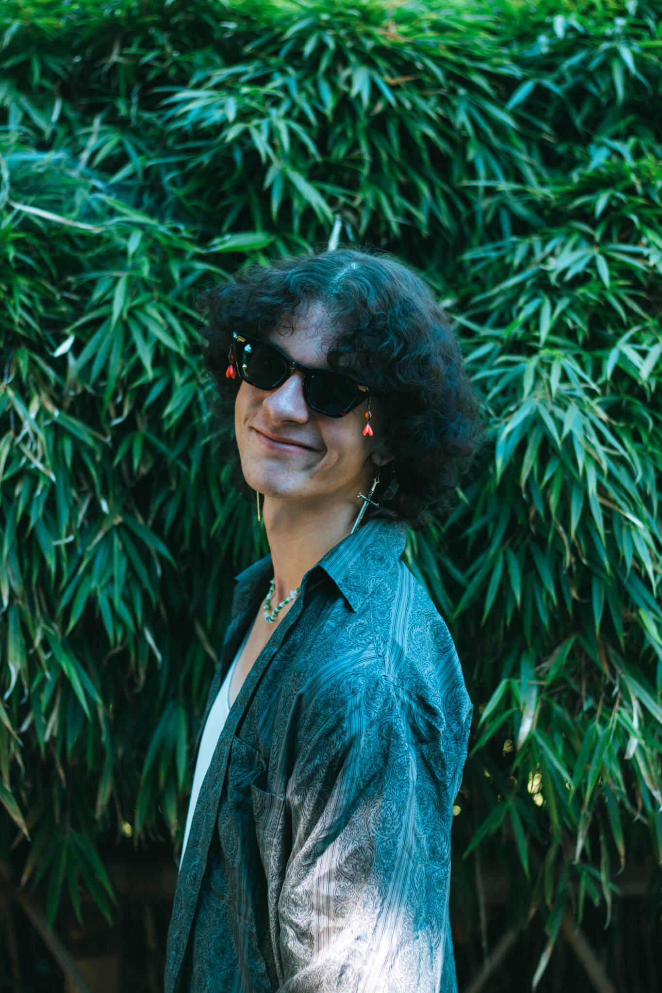A man wearing sunglasses and with curly hair poses in front of a vibrant green bush, showcasing a cheerful demeanor.