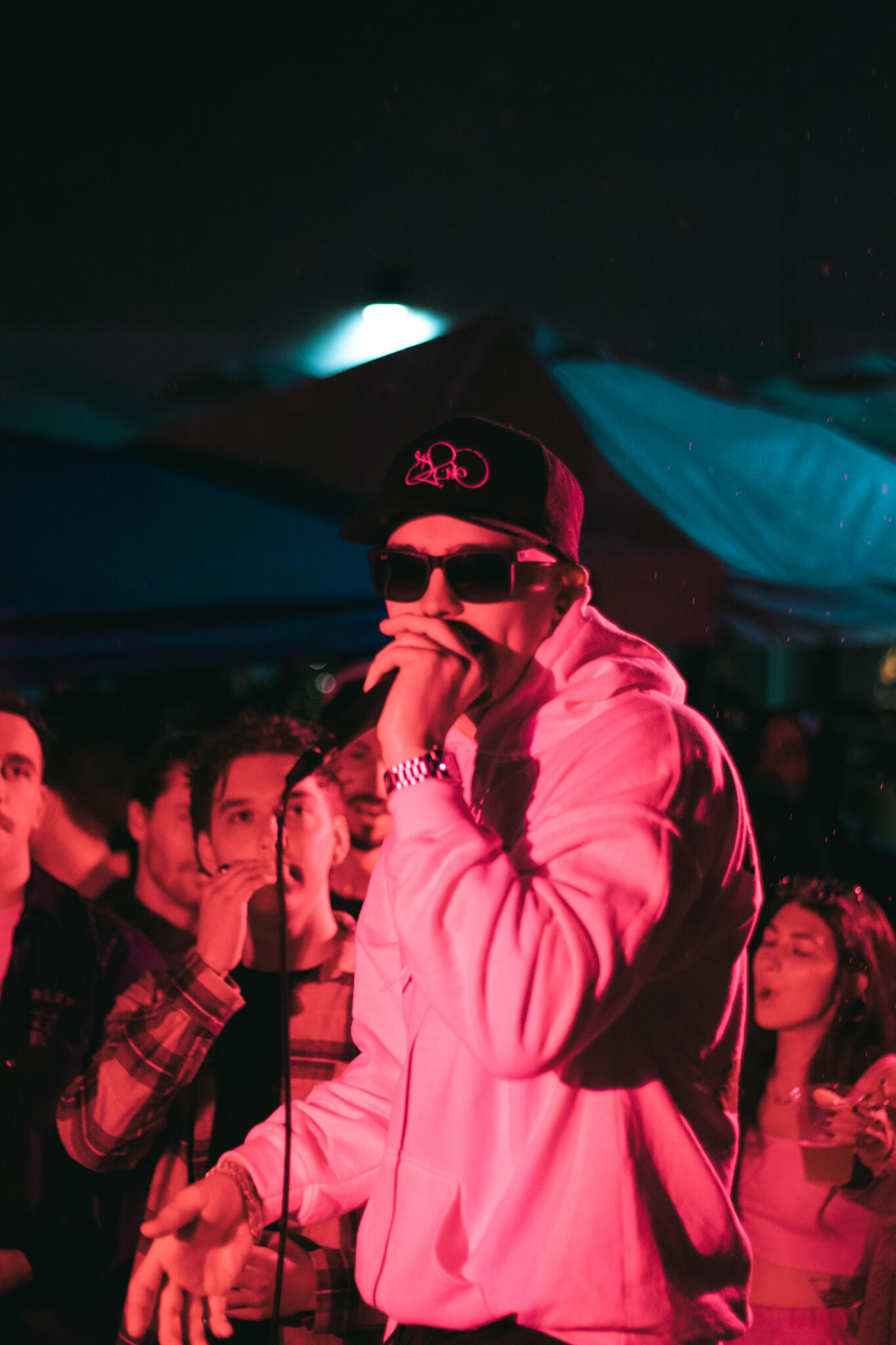 A man in a white hoodie illuminated by pink lighting and wearing sunglasses performs in front of a large audience in the rain while singing into a microphone.